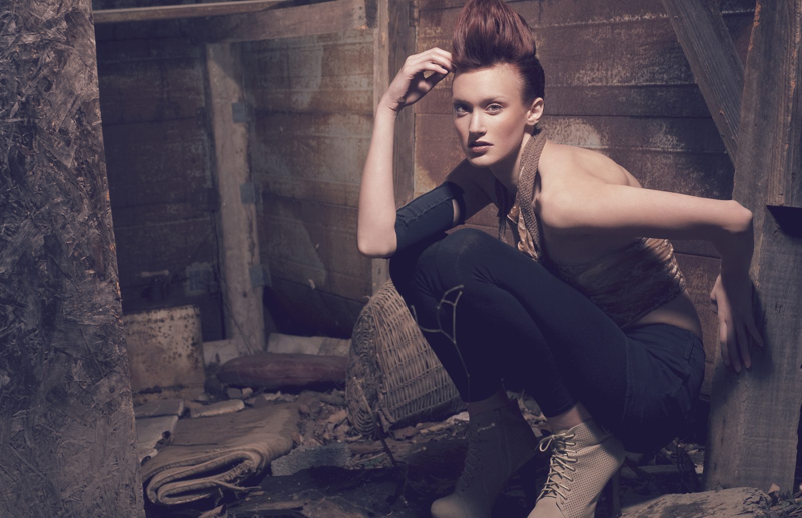 Model crouching in a wooden shed in lace-up boots, dark jeans and a cutout top with a black arm band