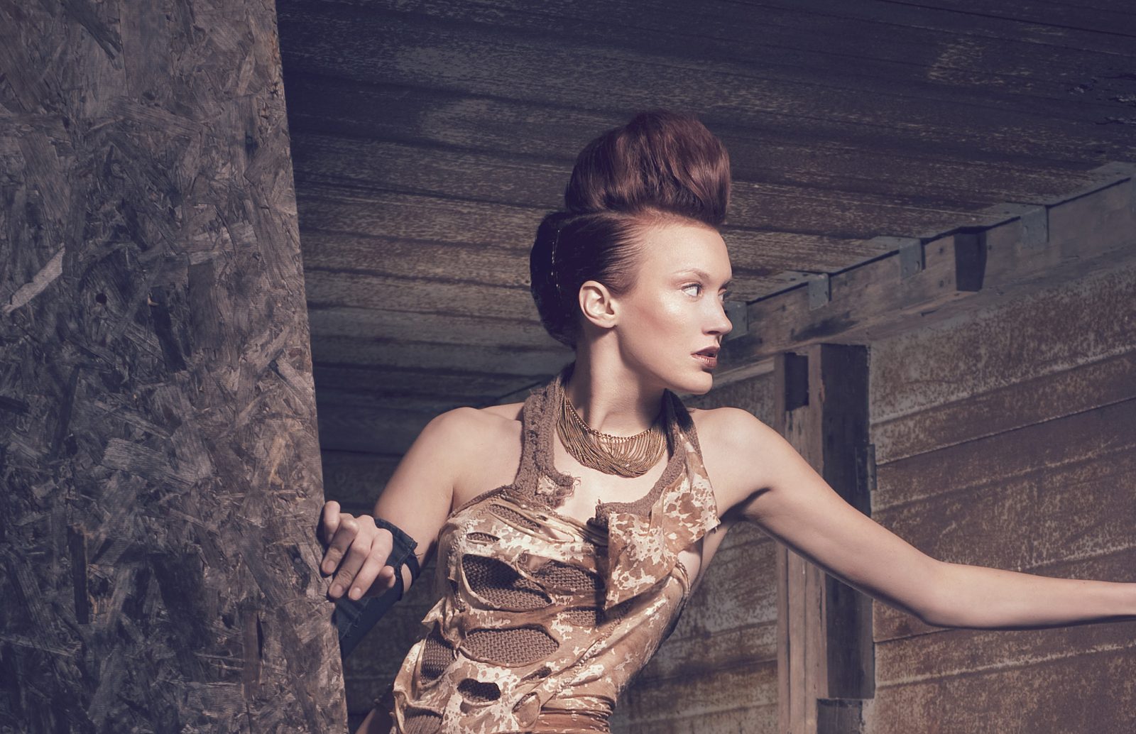 Model with a tall pompadour in a metallic bronze cutout top, bracing against a wooden post