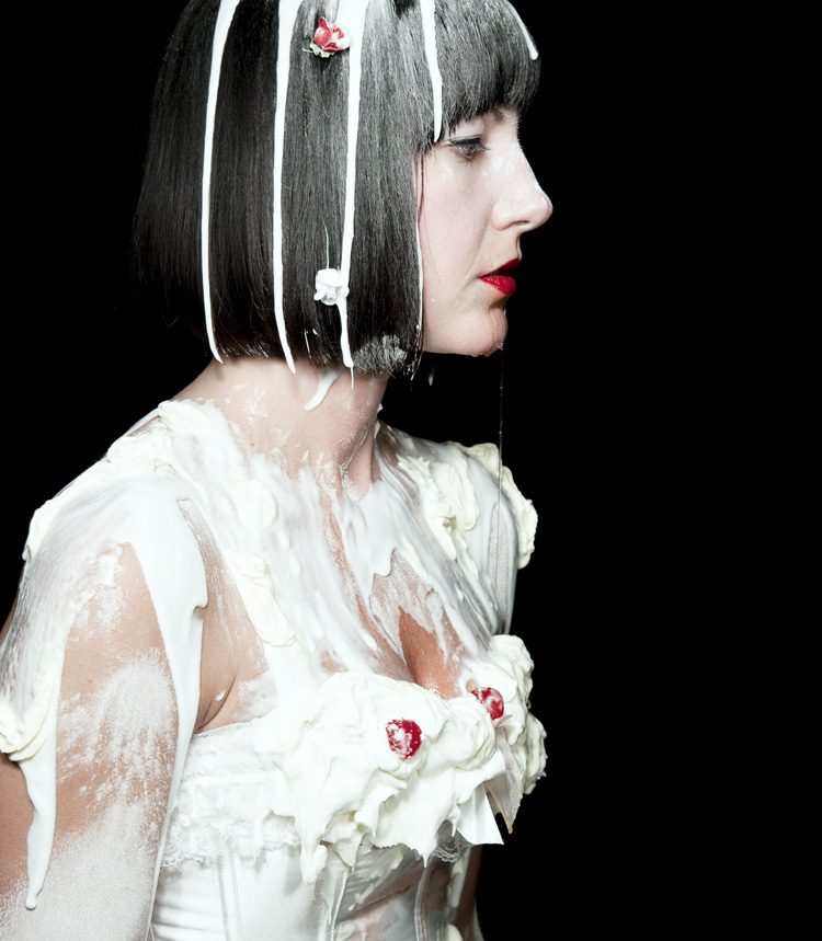 A woman with a sleek black bob and red lips in profile, her white corset and hair drenched in dripping whipped cream and topped with cherries