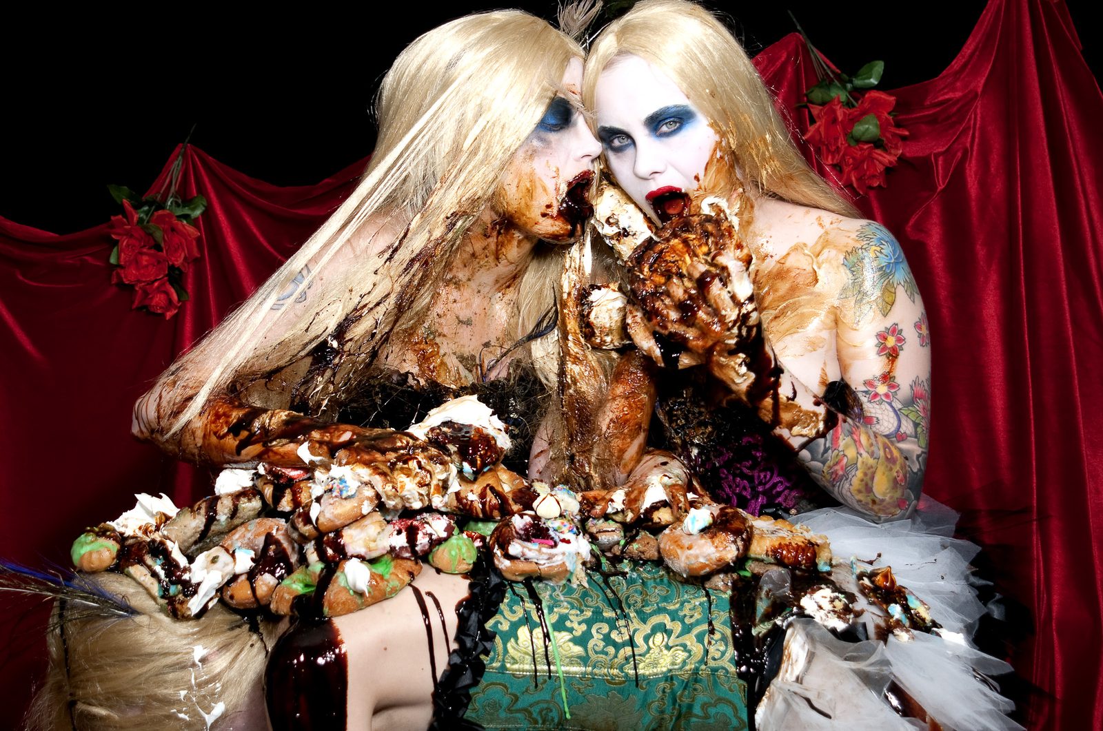 Two women in long blonde wigs, white face paint and blue eye makeup, smeared in chocolate syrup and frosting, devouring a heap of cakes and pastries in front of red velvet drapes and red roses