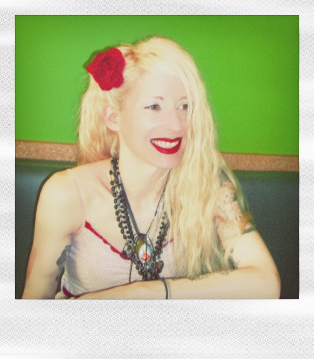 Polaroid portrait of Victoria F. Gaitán smiling, with long platinum hair, a red rose tucked behind her ear and layered necklaces