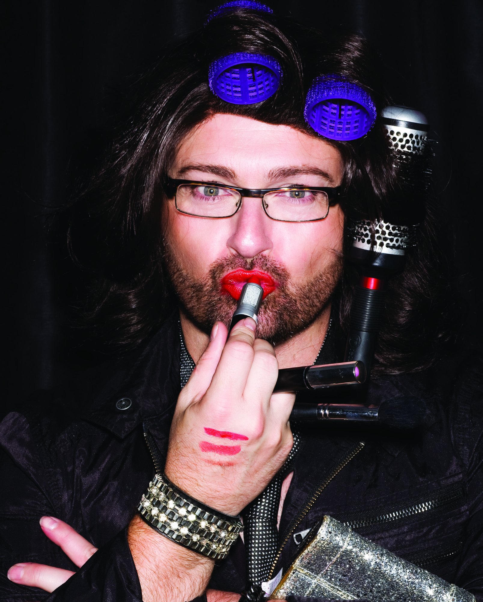 James Cornwell in a dark wig with purple rollers and glasses, applying red lipstick, holding a hair dryer and round brush