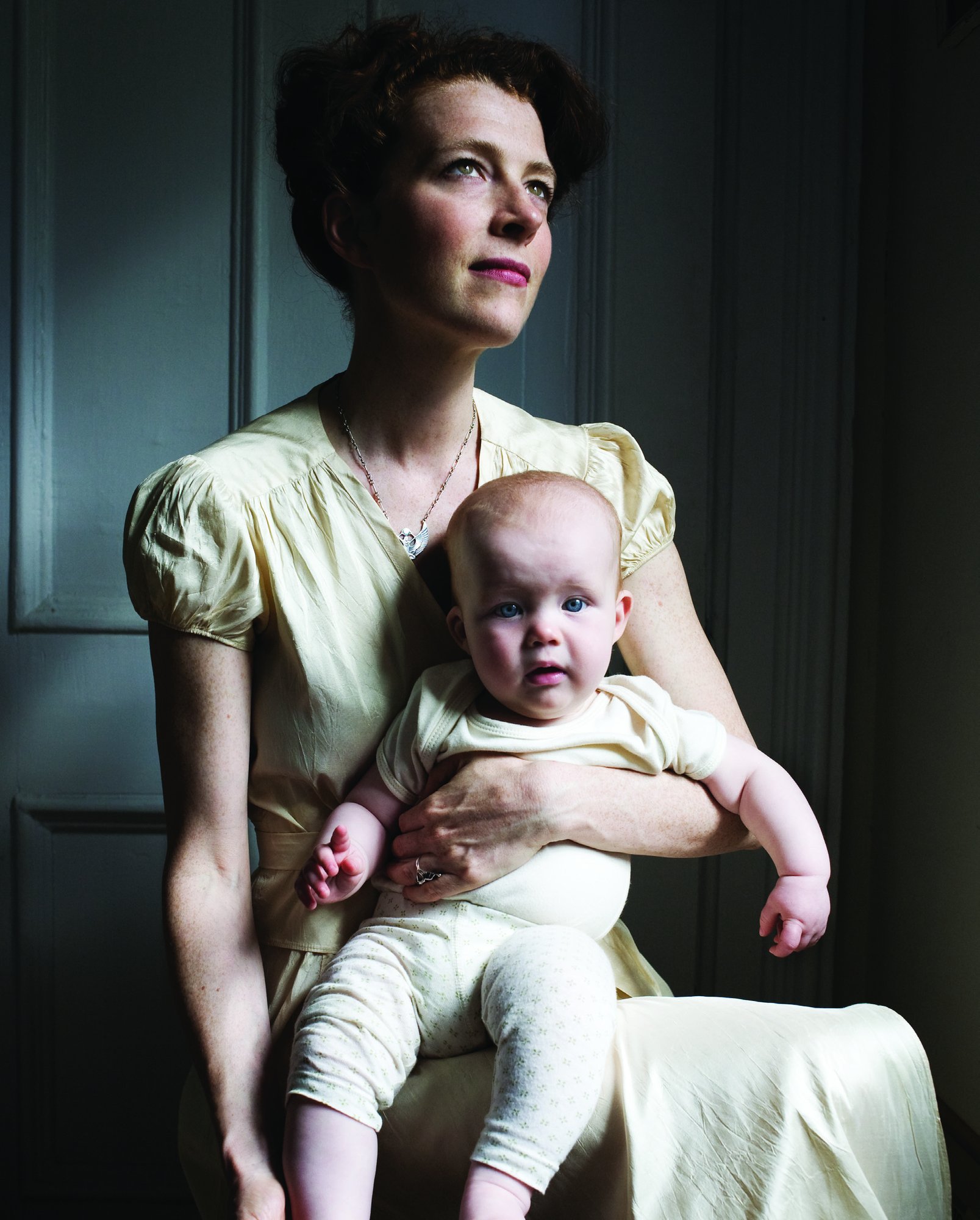 Melissa Auf der Maur in a cream vintage dress seated in soft window light, holding her baby daughter on her lap