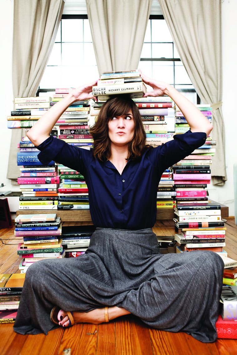 Svetlana Legetic sitting cross-legged on the floor surrounded by towering stacks of books, balancing more books on her head