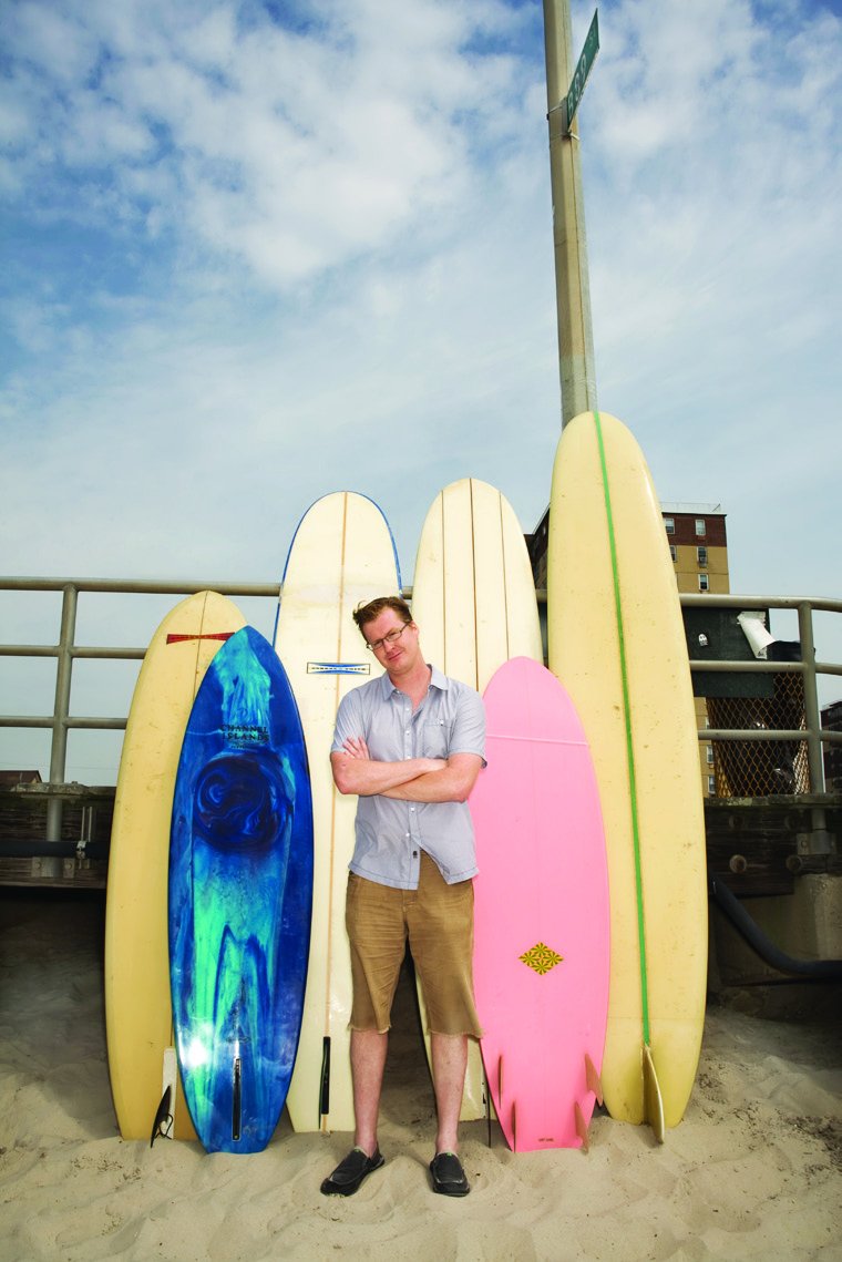 Kurt Braunohler standing on a beach with arms folded between a row of tall surfboards
