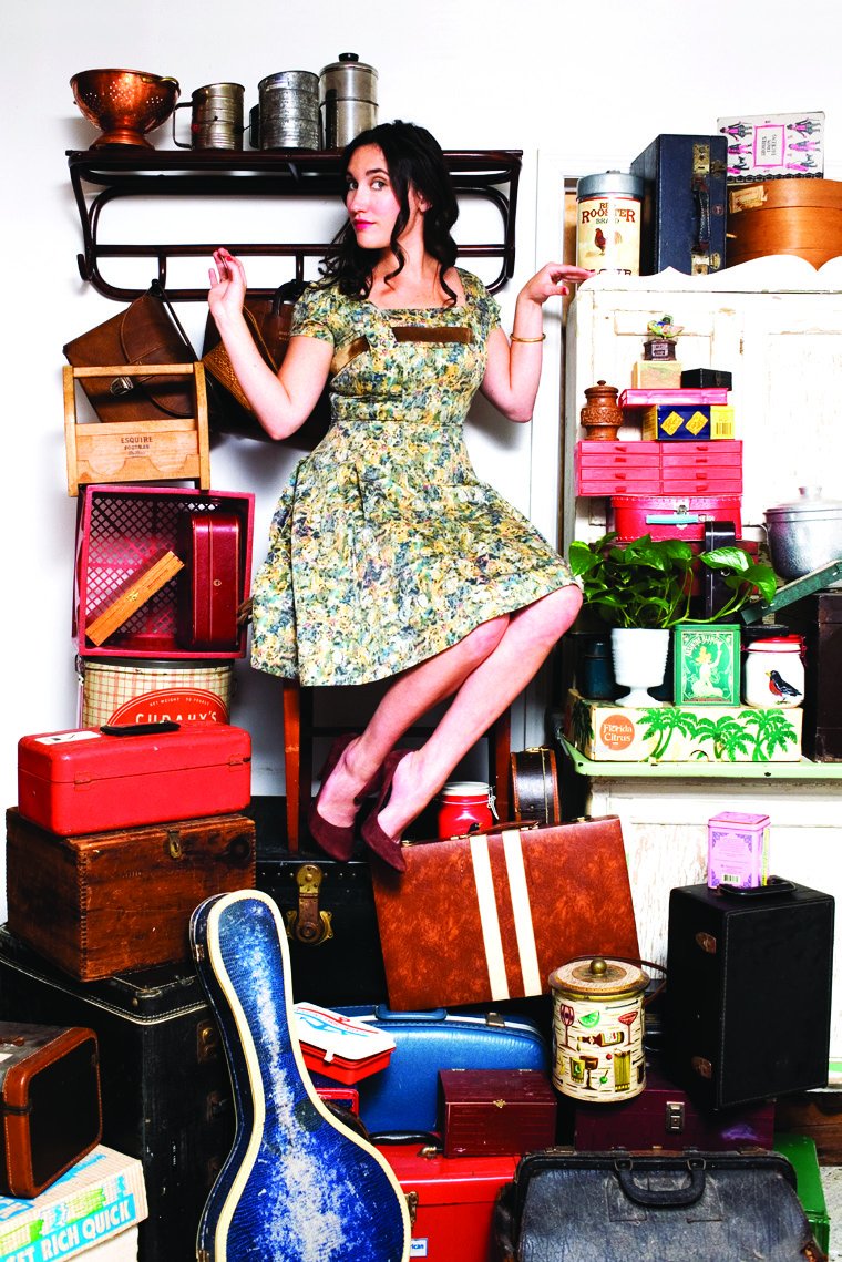 Susannah Hornsby in a floral dress perched atop a heap of vintage suitcases, tins, trunks and cases