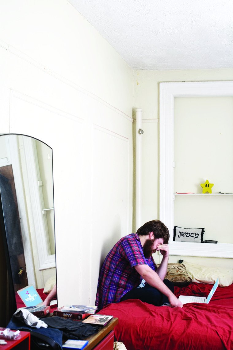 Dan St. Germain hunched on a red bed in a small apartment, looking at a laptop