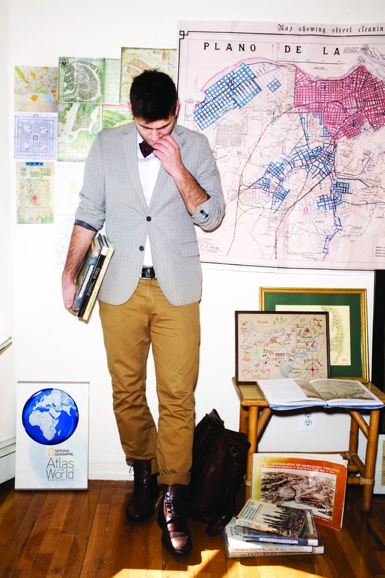 Dash Speaks in a gray blazer and bow tie standing in front of walls covered in vintage maps, beside a globe