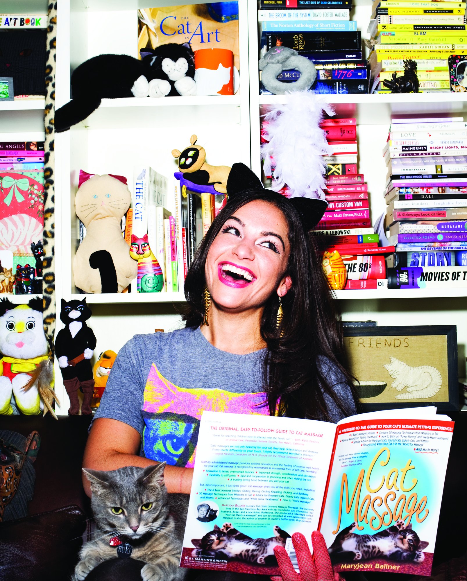 Saryn Chorney laughing in cat ears, holding a cat book and a gray cat, in front of shelves crowded with cat figurines and toys