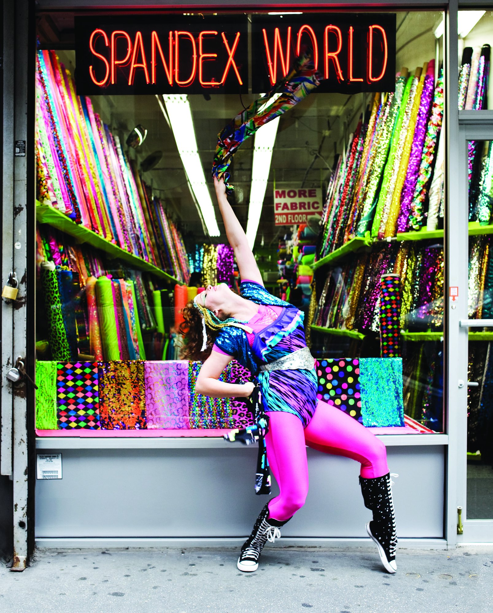 Adira Amram in hot pink leggings and a sequined top striking a high-kick pose outside the neon-signed Spandex World fabric store