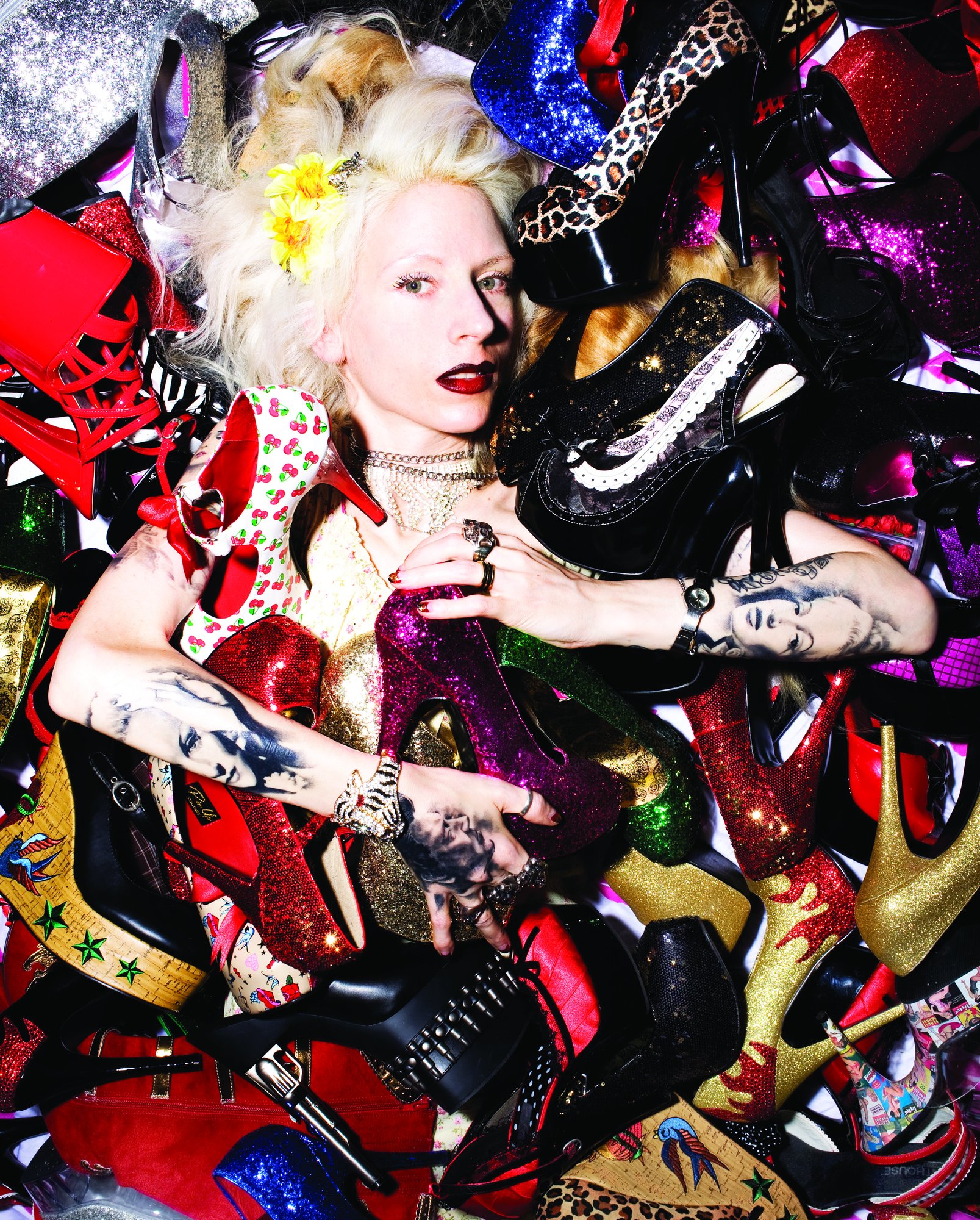 Victoria F. Gaitán with platinum hair and flowers, lying in a huge pile of glittering heels and boots