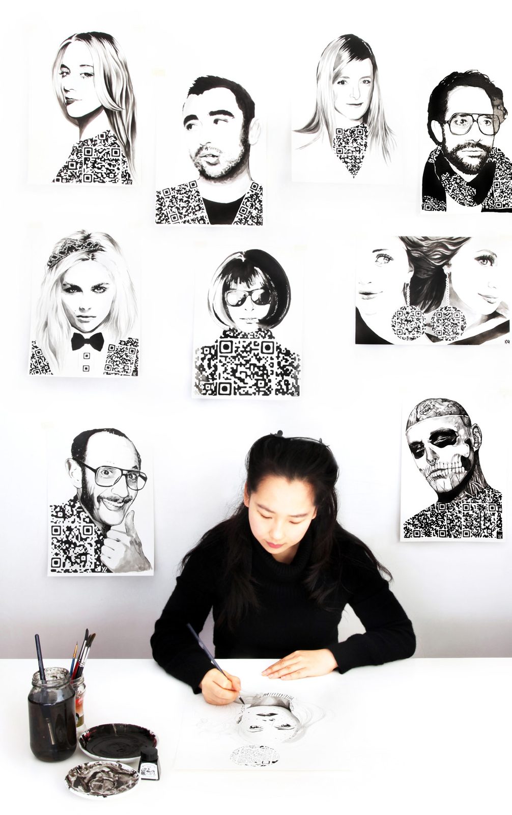 Yiying Lu sketching at a white desk beneath a wall of her black and white illustrated portraits