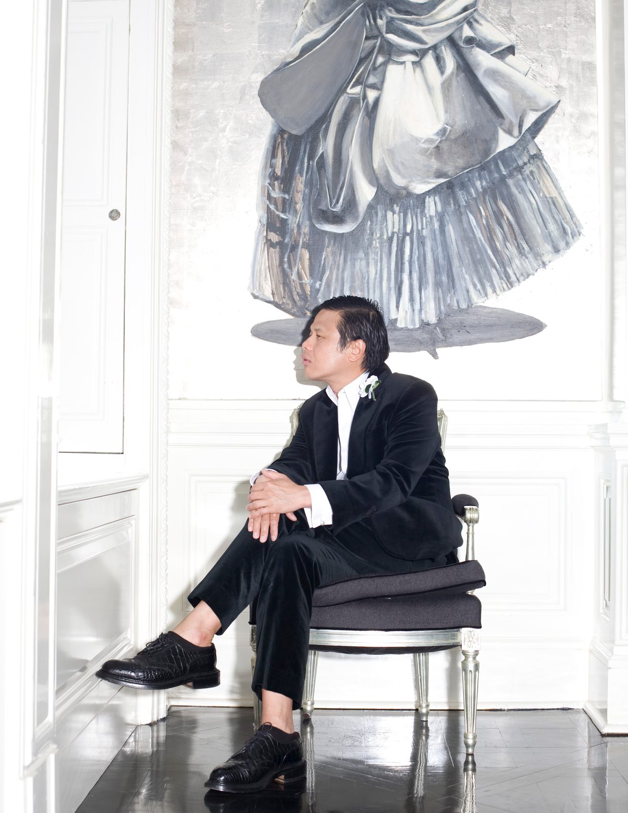 Zang Toi in a black suit seated on a chair in a white room beneath a large sculptural silver gown