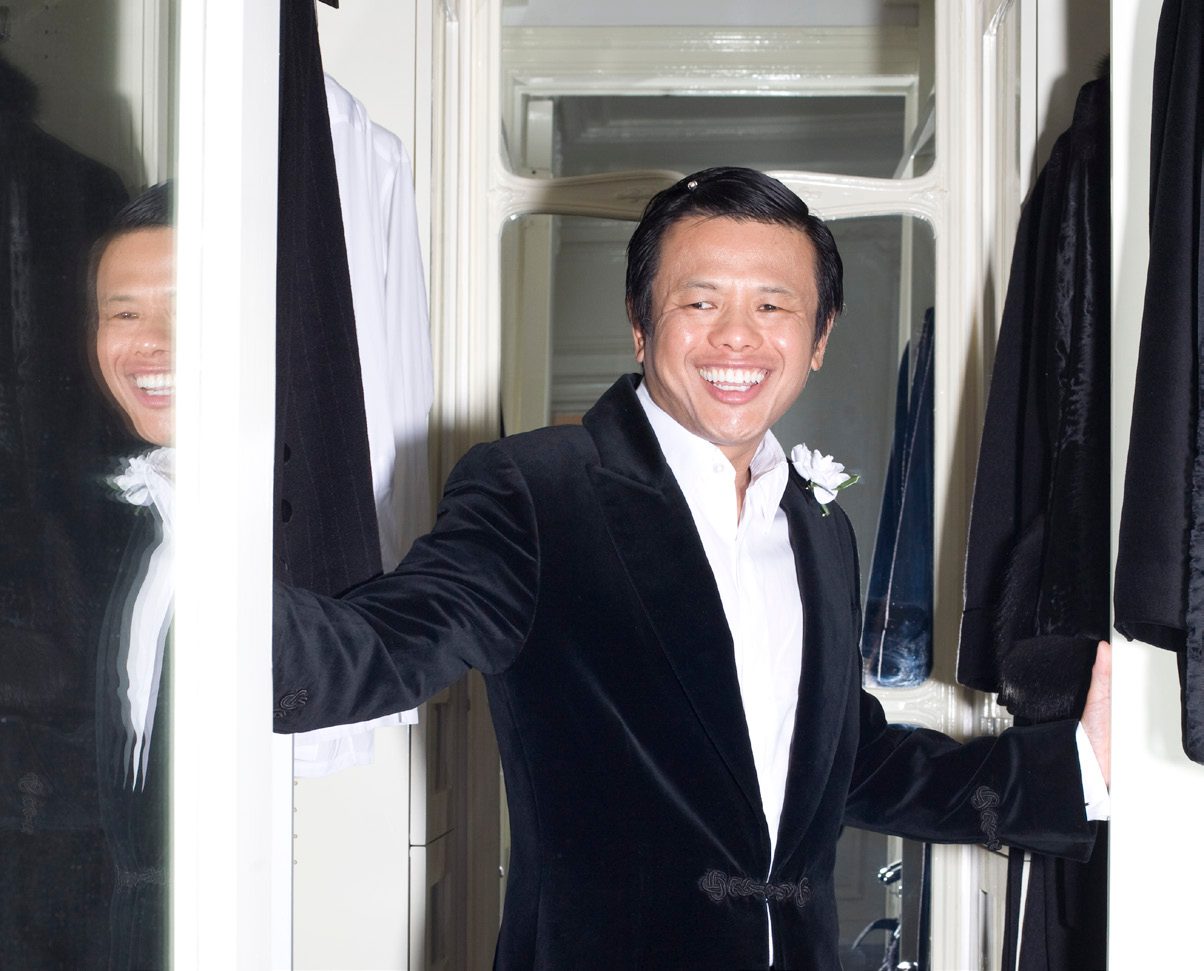 Zang Toi smiling in a black velvet tuxedo jacket with a white flower boutonniere, holding open a closet of garments